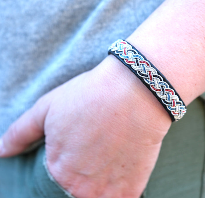 Handgjort armband i svart renskinn med färgstark tenntrådsfläta och renhornsknapp, inspirerat av Gammelstad Kyrkstad och dess historiska miljö.
