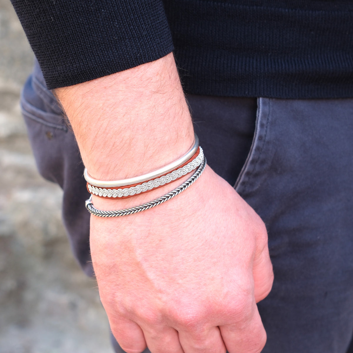 Närbild på mans handled med handflätat tennarmband i silverfärgad tenntråd på ljusbrunt renskinn, kombinerat med två silverfärgade armband