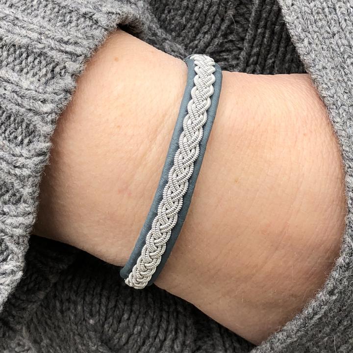 Närbild på handflätat tennarmband Arctic med silverfärgad tenntråd på petrolfärgat renskinn, buret på handleden tillsammans med en grå stickad tröja. handgjord av Arctic Collection i Gammelstad
