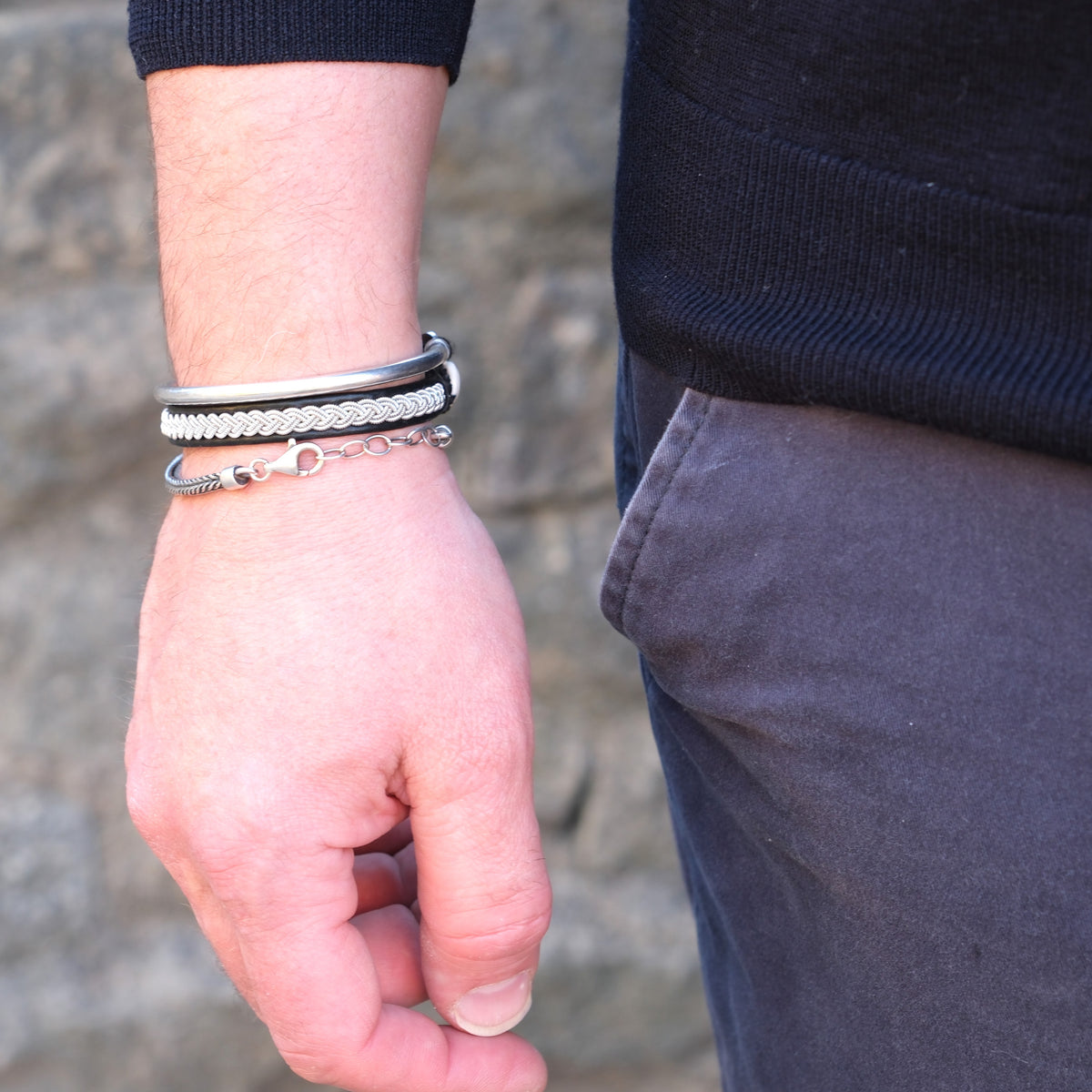 Närbild på mans handled med handflätat tennarmband Arctic på svart renskinn, kombinerat med silverfärgade armband från Arctic Collection.