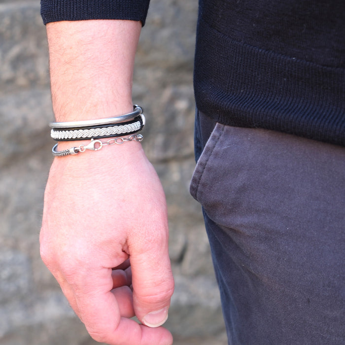 Närbild på mans handled med handflätat tennarmband Arctic på svart renskinn, kombinerat med silverfärgade armband från Arctic Collection.