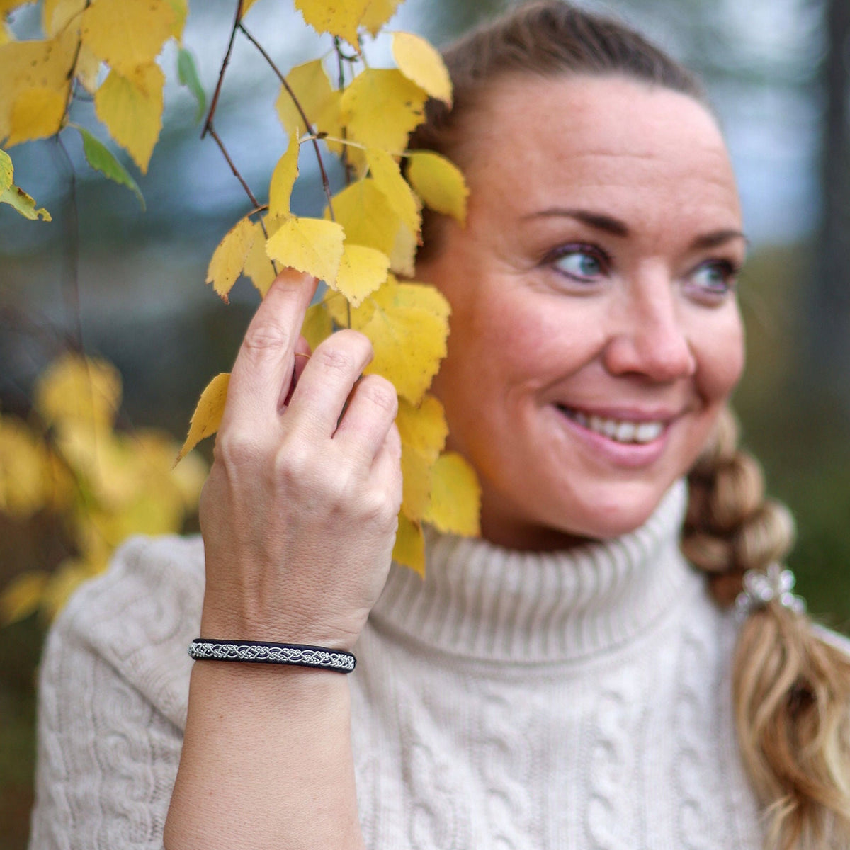 En kvinna i naturvit stickad tröja bär tennarmbandet Aurora från Arctic Collection, med silverfärgad tenntrådsfläta på svart renskinn. Hon står utomhus bland gula höstlöv.