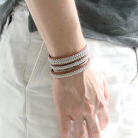 Närbild på tre tennarmband Is i cognacsfärgat renskinn med handflätad silverfärgad tenntråd från Arctic Collection. Armbanden bärs tillsammans på handleden till ljusa byxor