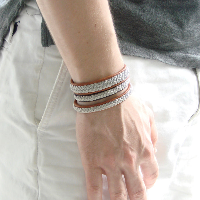 Närbild på tre tennarmband Is i cognacsfärgat renskinn med handflätad silverfärgad tenntråd från Arctic Collection. Armbanden bärs tillsammans på handleden till ljusa byxor