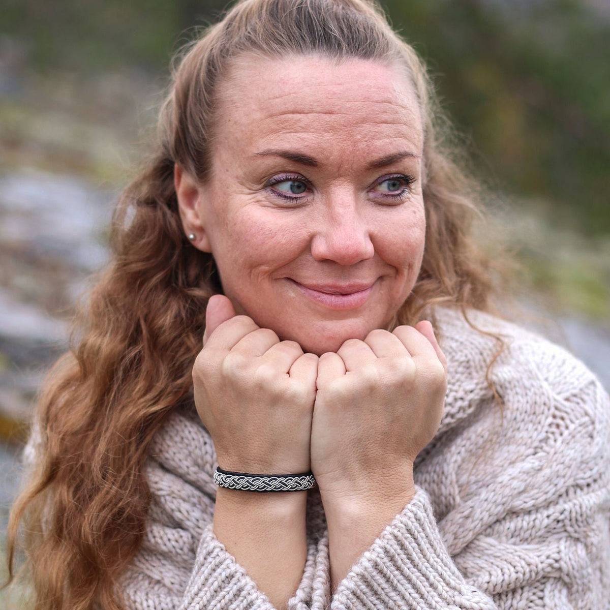 Leende kvinna i ljus stickad ulltröja bär det handflätade tennarmbandet Kust på svart renskinn, sittande på en mossbeklädd klippa i nordisk skogsmiljö.