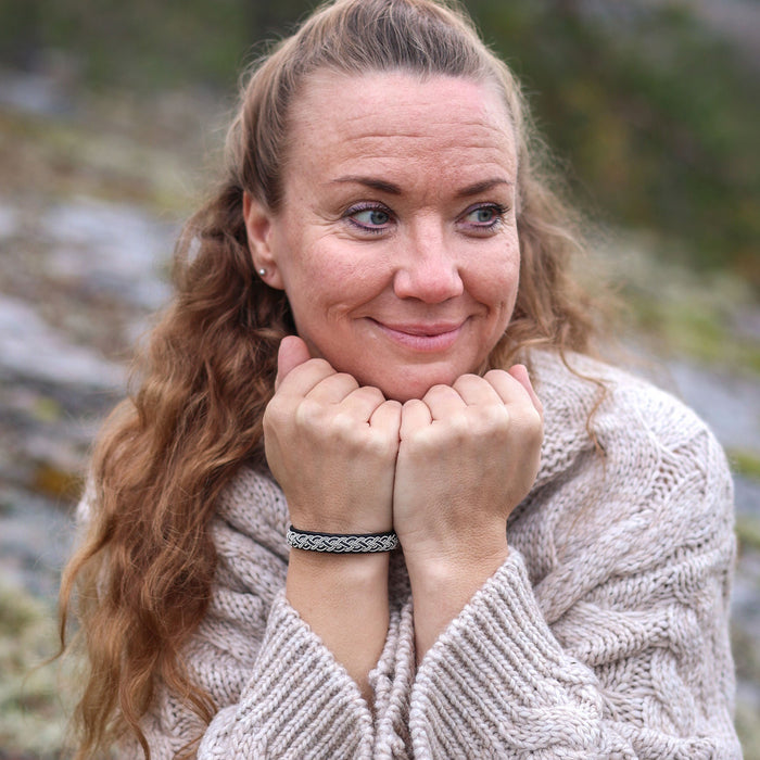Leende kvinna i ljus stickad ulltröja bär det handflätade tennarmbandet Kust på svart renskinn, sittande på en mossbeklädd klippa i nordisk skogsmiljö.