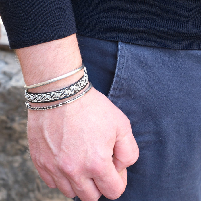 Närbild på mans handled med det handflätade tennarmbandet Kust på svart renskinn, kombinerat med två silverfärgade armband i olika mönster.