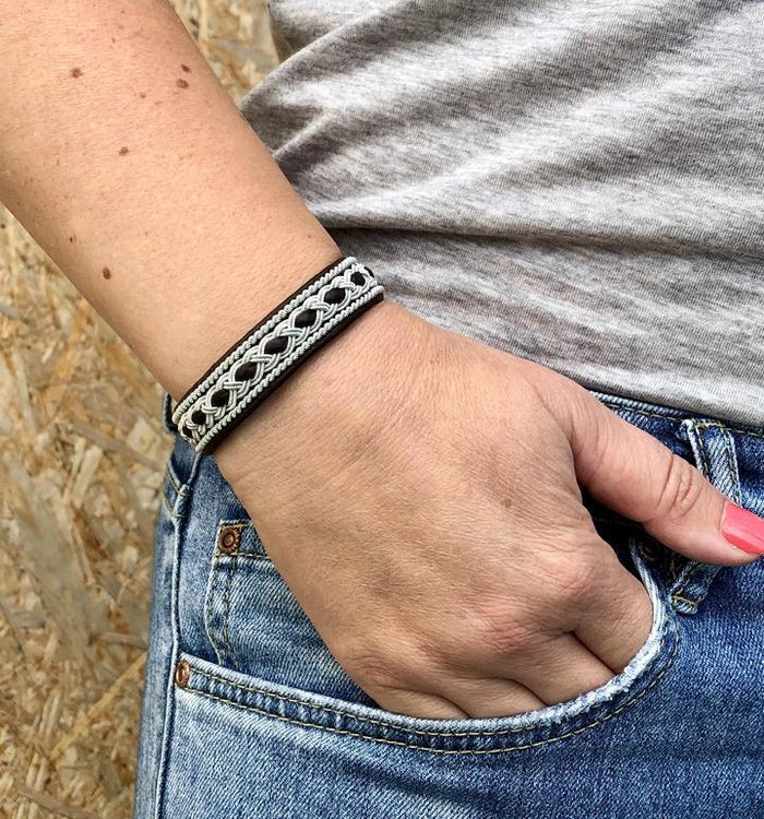 Tennarmband Kyla från Arctic Collection i svart renskinn med handflätad tenntråd. Närbild på armbandet runt en kvinnas handled, med handen i jeansfickan. 