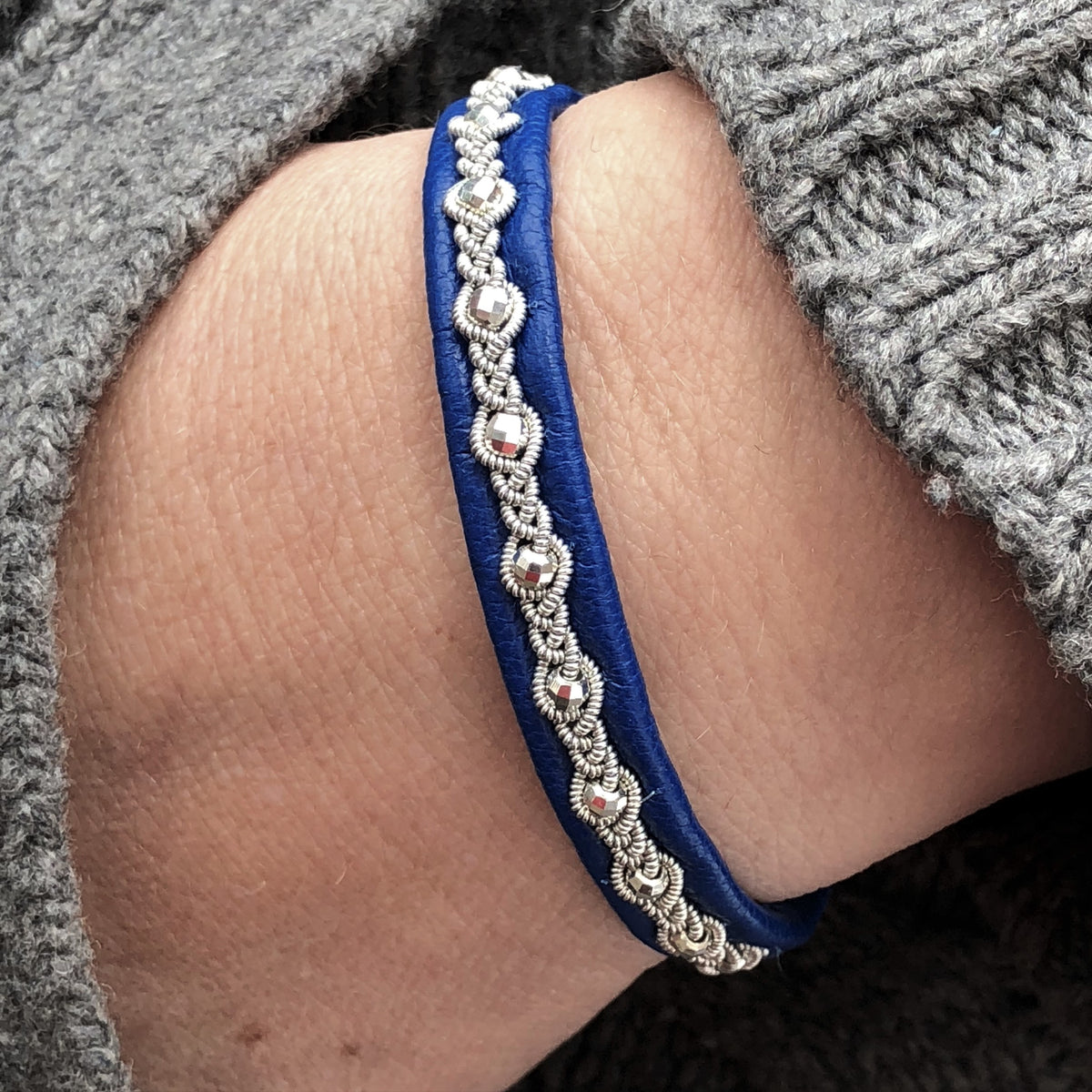 Tennarmband Midnattssol från Arctic Collection i mörkblått renskinn med handflätad tenntråd. Armbandet sitter på en handled med gråstickad tröja.