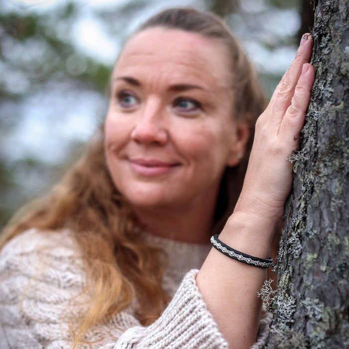 Tennarmband Midnattssol från Arctic Collection i svart renskinn med handflätad tenntråd. En kvinna i naturvit stickad tröja bär armbandet med handen mot en trädstam. Kopiera