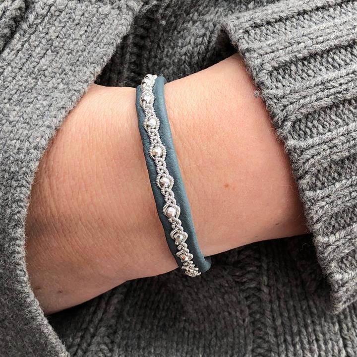 Tennarmband Midnattssol från Arctic Collection i petrol renskinn med handflätad tenntråd. Armbandet sitter på en handled med gråstickad tröja.