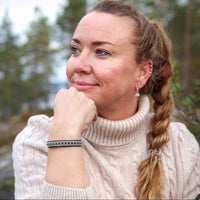 En kvinna i naturvit stickad tröja bär tennarmbandet Nordvind från Arctic Collection, med silverfärgad tenntrådsfläta på svart renskinn. Hon sitter utomhus i skogsmiljö med hakan vilande mot handen