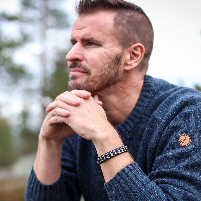 Man i mörkblå stickad tröja bär det handflätade tennarmbandet NORR med silverfärgad tennfläta på svart renskinn från Arctic Collection, fotograferad utomhus.