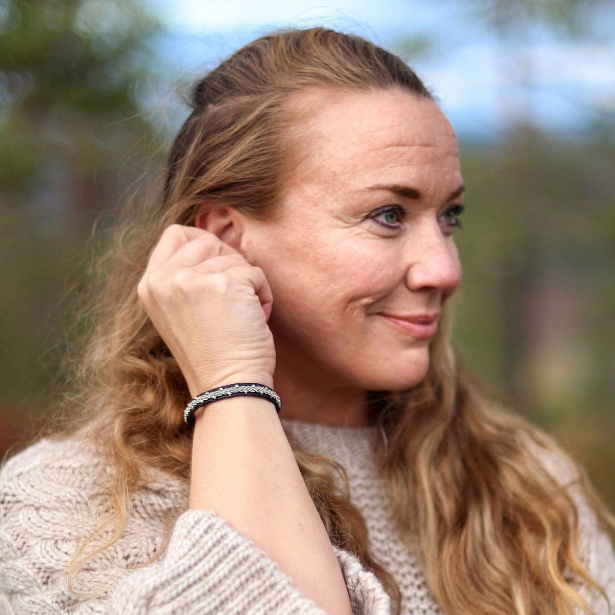 Tennarmband Polarnatt från Arctic Collection med handflätad tenntråd på en kvinnas handled. Kvinnan bär en naturvit stickad tröja och står utomhus i nordisk natur. Kopiera