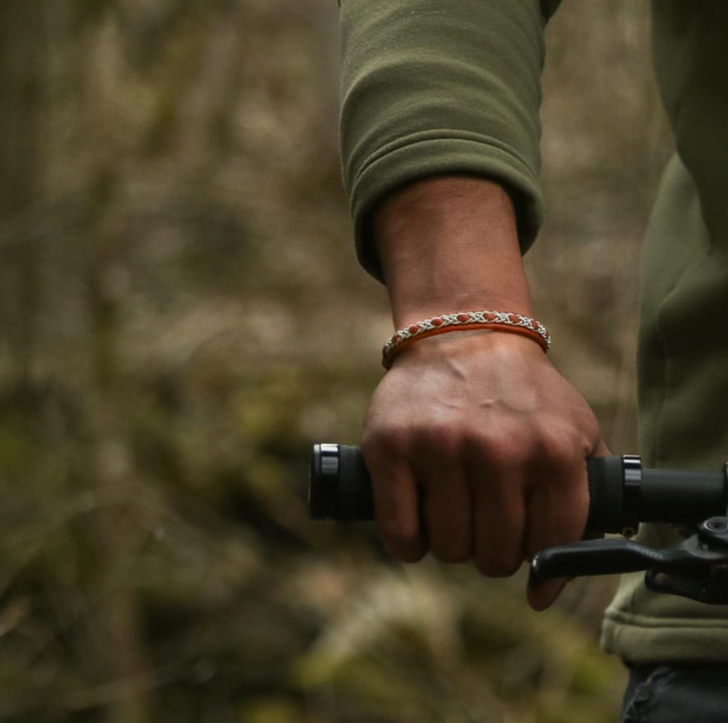 Tennarmbandet Snö från Arctic Collection i ljusbrunt renskinn med silverfärgad tenntrådsfläta, på handleden hos en person som håller i ett cykelstyre utomhus.