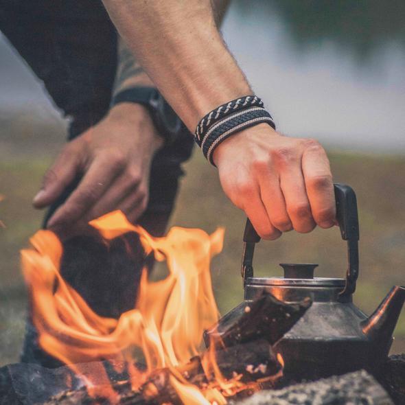 Närbild på mans handled med svarta handflätade tennarmband från Arctic Collection, medan han håller en kaffepanna över en öppen eld i nordisk natur.