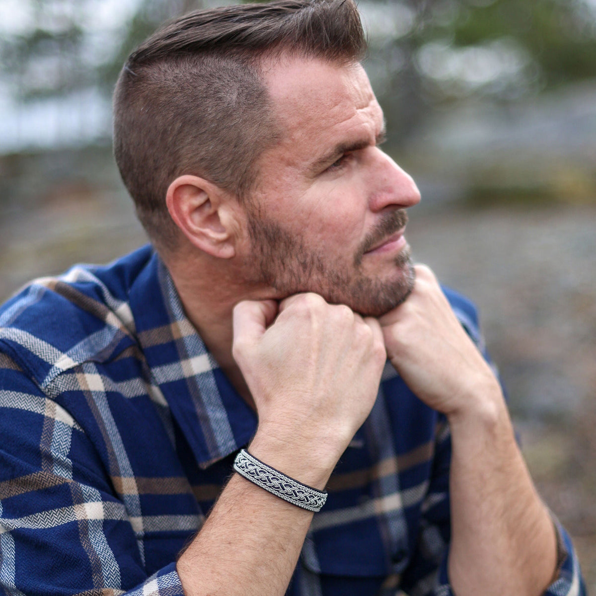 Man i blå rutig skjorta och jeans bär det handflätade tennarmbandet Taiga på svart renskinn från Arctic Collection, sittande utomhus i nordisk natur.