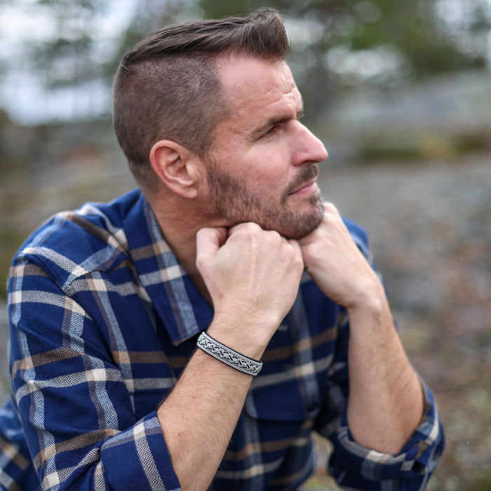 Man i blå rutig skjorta och jeans bär det handflätade tennarmbandet Taiga på svart renskinn från Arctic Collection, sittande utomhus i nordisk natur.