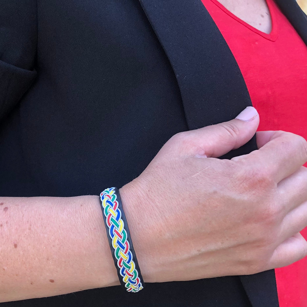 Tennarmband Tradition från Arctic Collection i svart renskinn med färgglad fläta av tenntråd. Armbandet bärs på en kvinnas handled tillsammans med en svart kavaj och röd topp. 