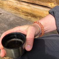 Närbild på en handled med det ljusbruna tennarmbandet Tundra från Arctic Collection, handgjort i renskinn med silverfärgad tenntrådsfläta. Personen håller en kaffekopp utomhus