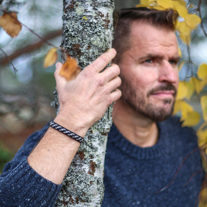 Man med svart tennarmband Tundra från Arctic Collection, med silverfärgad tenntrådsfläta på renskinn. Han står vid ett träd med gula höstlöv
