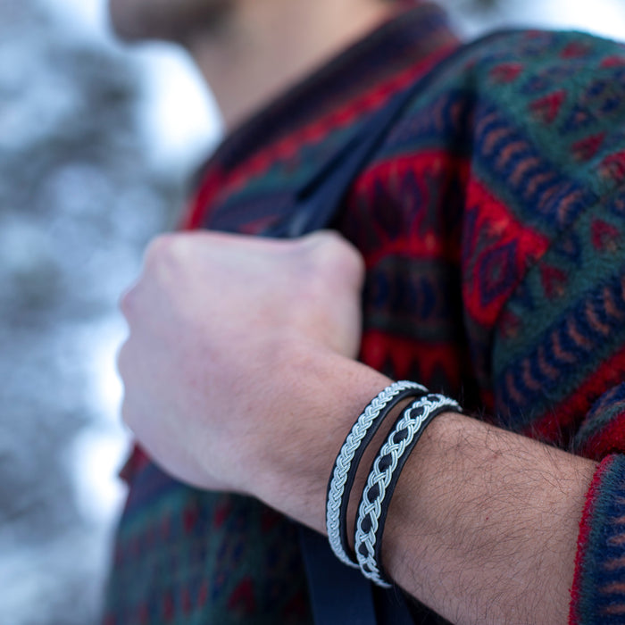 Närbild på mans handled med två handflätade tennarmband på svart renskinn, varav modellen Vildmark från Arctic Collection, tillsammans med en färgglad ulltröja i vintrig natur.