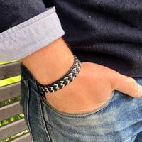 Närbild på tennarmbandet NORR med bred svart läderrem och handflätat sicksackmönster i silverfärgad tenntråd, buret av en man med handen i jeansfickan.